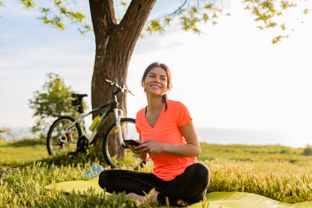 smiling beautiful woman holding phone doing sports in morning in park nature on mat wearing pink shirt fitness outfit, happy healthy lifestyle, music in earphones, bicycle on background